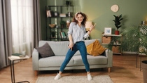 Happy joyful young Caucasian beautiful woman in positive mood dancing moving rhythmically and jumping while listening to music song in headphones on smartphone, having fun in room, leisure concept