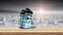 Light blue ski boot on a wooden surface with a snowy mountain landscape in the background. 

