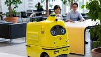 MobiPower, a bright yellow autonomous delivery robot, operating inside a modern indoor workspace with tables, seating areas, and plants. MobiPower, a bright yellow autonomous delivery robot, operating inside a modern indoor workspace with tables, seating areas, and plants.