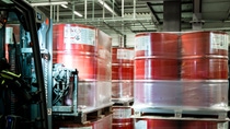 Forklift transporting pallets of red industrial drums wrapped in plastic inside a warehouse.