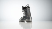 Black ski boot standing on a reflective surface against a light gray background. 
