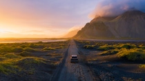 A single car drives along a lonely gravel road in golden sunlight, surrounded by mountains and open countryside