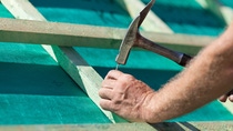 Hand using a hammer to fasten a nail into a wooden frame on a construction surface.