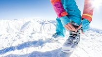 Person fastening a ski boot in deep snow with snow-covered mountains in the background.
