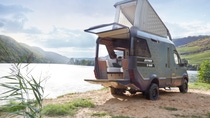 Hymer VisionVenture camper van parked beside a scenic lakeside landscape with open rear access and an elevated pop‑up roof. Hymer VisionVenture camper van parked beside a scenic lakeside landscape with open rear access and an elevated pop‑up roof.