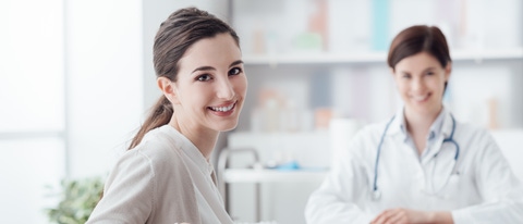 Smiling patient receiving a medical consultation and looking at camera, the female doctor is sitting at desk on the background Smiling patient receiving a medical consultation and looking at camera, the female doctor is sitting at desk on the background
