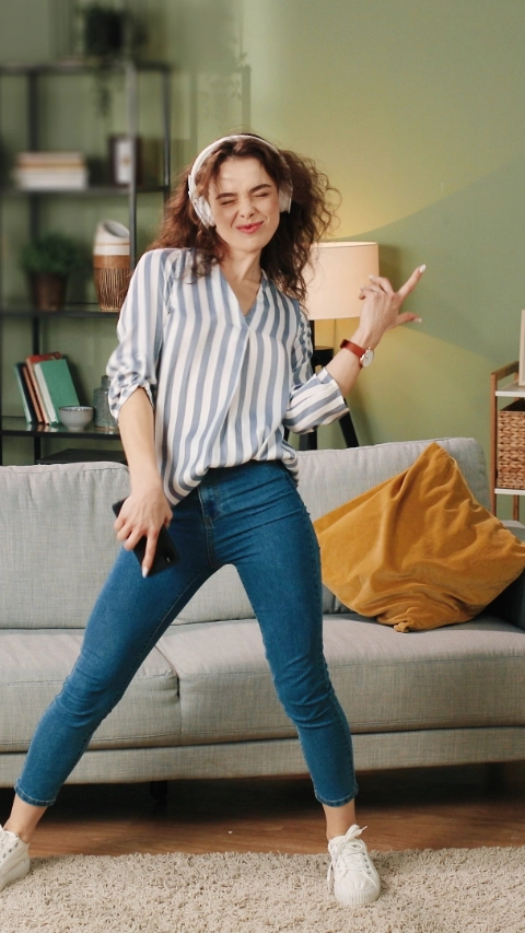 Happy joyful young Caucasian beautiful woman in positive mood dancing moving rhythmically and jumping while listening to music song in headphones on smartphone, having fun in room, leisure concept