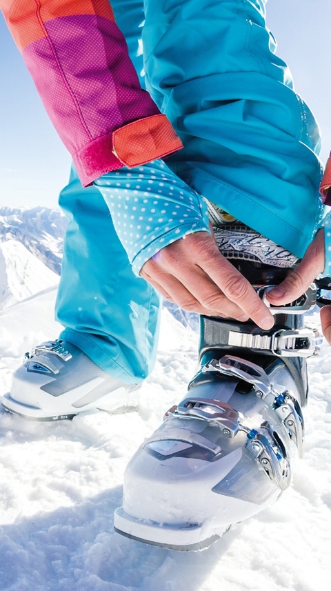 Person fastening a ski boot on a snowy mountain slope, with snow-covered peaks in the background.