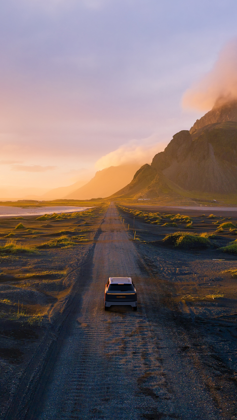 A single car drives along a lonely gravel road in golden sunlight, surrounded by mountains and open countryside